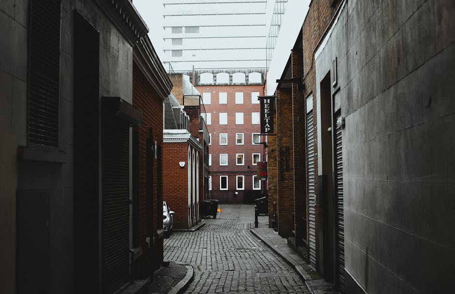 A narrow urban street scene showing a small, sloping tarmac driveway between two buildings. On the left, there are older, brick-built structures with several windows, some with external air conditioni