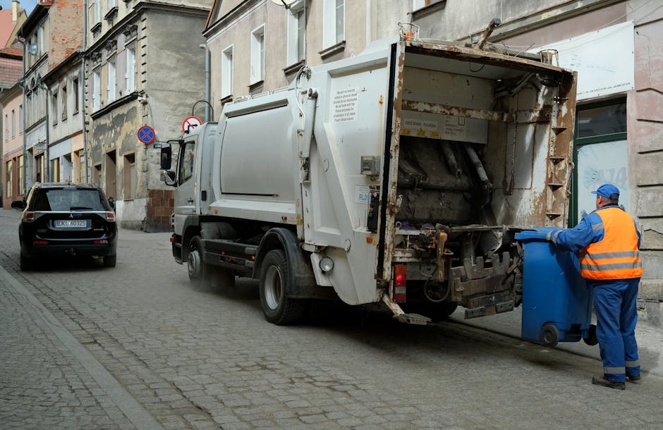 A red metal waste skip with visible rust and chipped paint sits on a paved area between two concrete or stone walls in an urban outdoor setting. The large container has a sliding door on its side, and