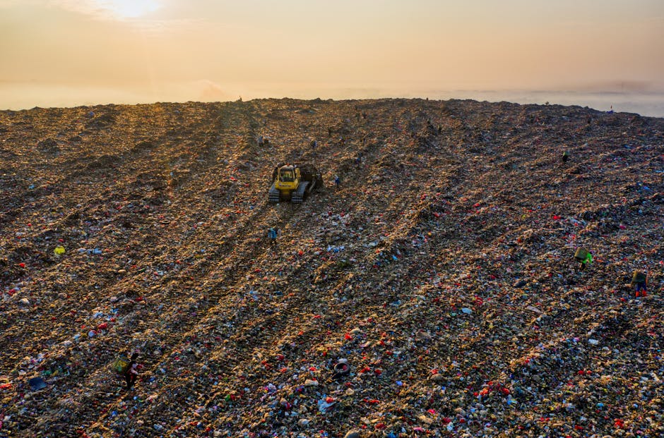 A large open landfill site during daylight hours, featuring multiple yellow excavators actively moving heaps of mixed waste, primarily consisting of plastic bags, cardboard, fabric, and other refuse m